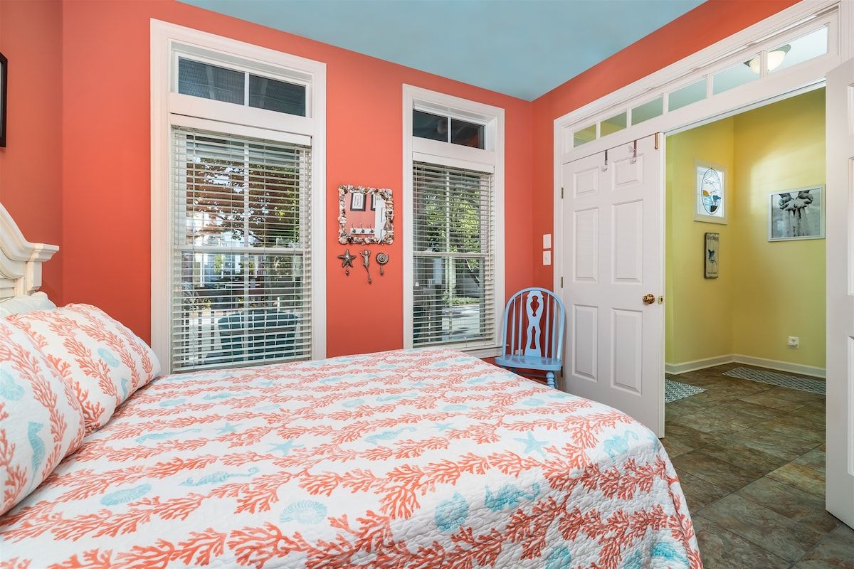 Un dormitorio con cama, dos ventanas con persianas, una puerta y un pasillo con otra puerta.