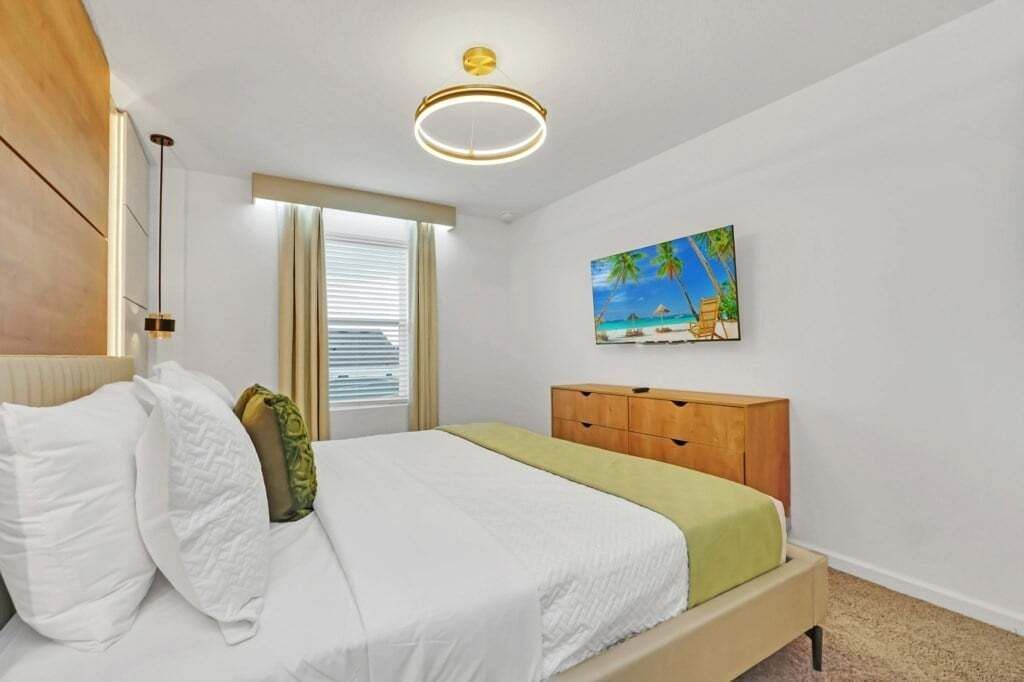 Un dormitorio con cama, cabecera de madera, mesita de noche, ventana con persianas y un cuadro de una playa, además de una lámpara de techo moderna.