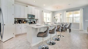 A modern kitchen with white cabinets, a central island with bar stools, a microwave, and a dining area with a table and chairs. - Orlando Newest Resort Villa With Pool 1791spp 4 Bedroom Home (Kissimmee)