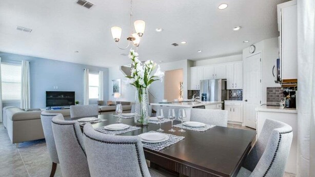 A modern kitchen with white cabinets, a dining area with a dark wooden table, and a living room with a sofa and TV. - Orlando Newest Resort Villa With Pool 1791spp 4 Bedroom Home (Kissimmee)
