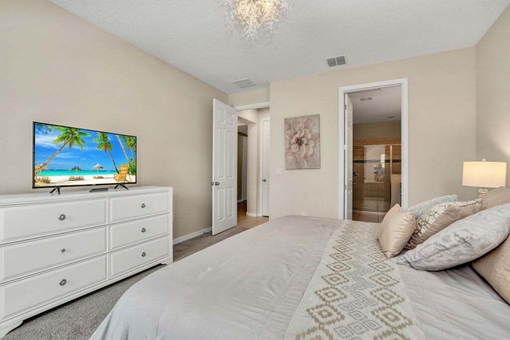 A bedroom with a bed, a TV on a dresser, a door leading to a bathroom, and a painting on the wall.