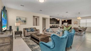 A spacious living room with a large TV, two sofas, a coffee table, and a dining area with a chandelier. - 1513nassau 5 Bedroom Villa (Kissimmee)