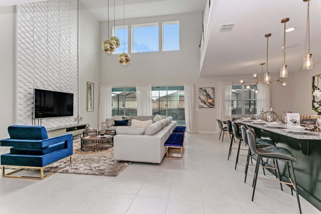 A spacious living area with a large flat-screen TV, a white sofa, a blue accent chair, and a dining area with a bar.