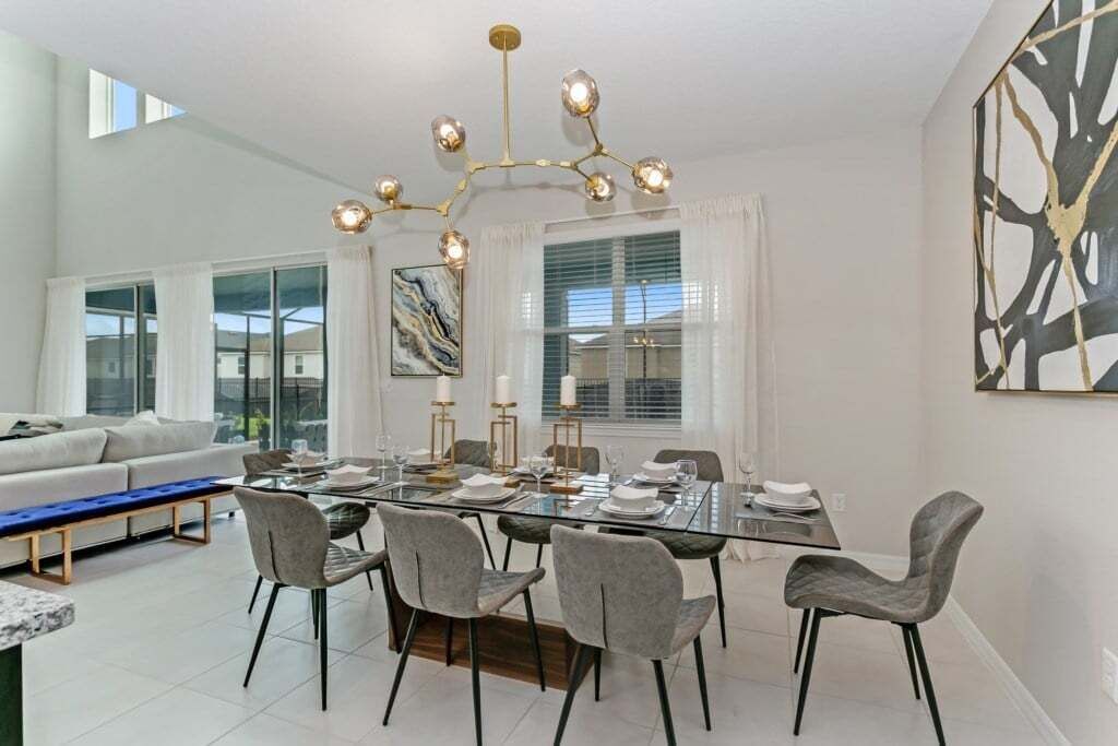 A modern dining area with a glass table, grey upholstered chairs, and a chandelier.
