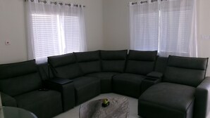 A modern living room with a sectional sofa, a glass coffee table, and two windows with sheer curtains.