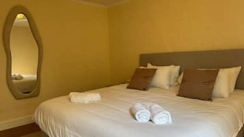 A neatly made bed with white linens and brown pillows, a large oval mirror on the wall, and a plain yellow wall.