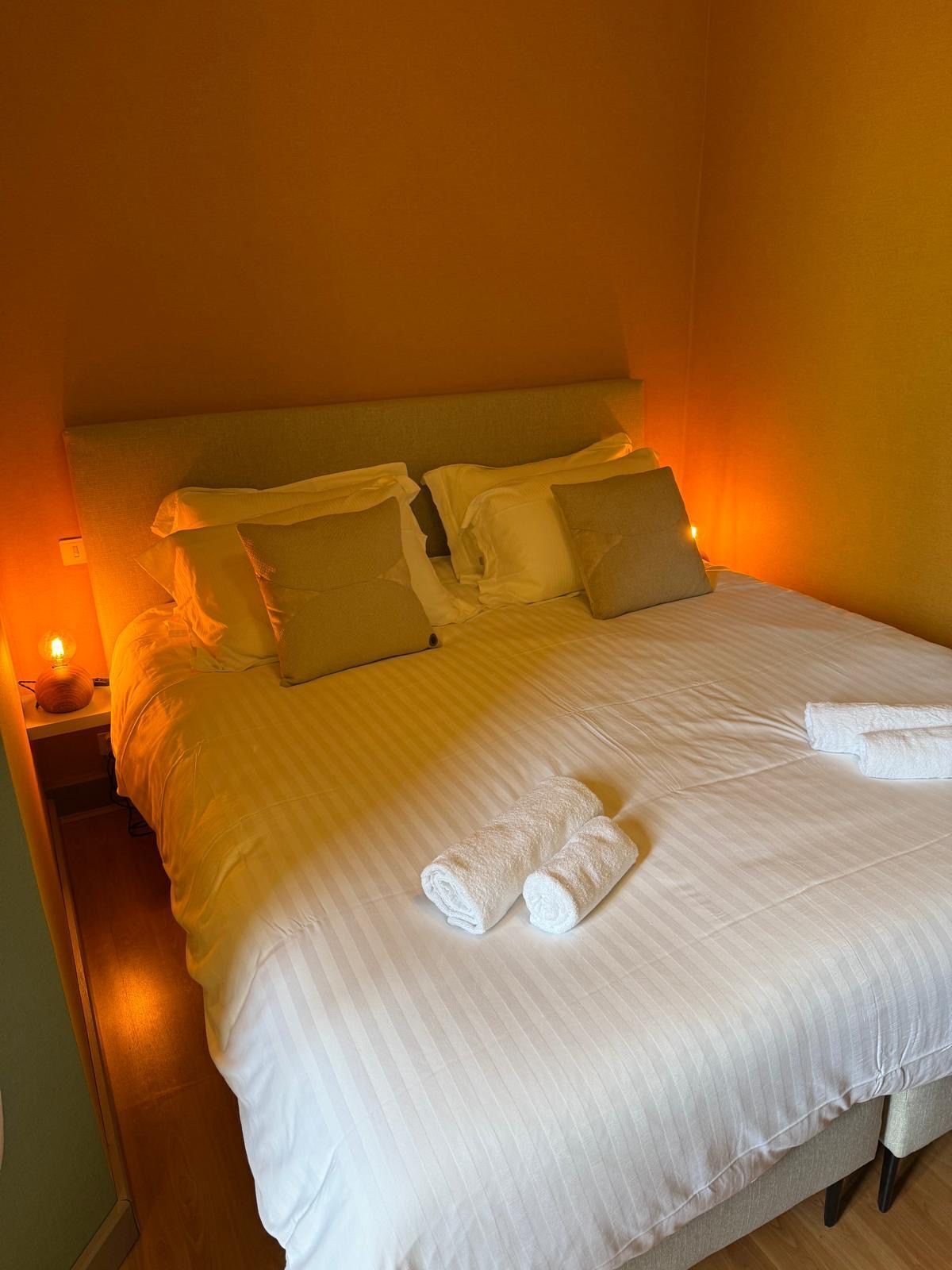 Una cama bien hecha con sábanas blancas, dos almohadas y toallas enrolladas sobre un suelo de madera.