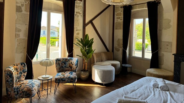 A hotel room with a large bed, two armchairs, a small table, a potted plant, and a view of the outside.