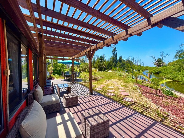 A covered outdoor patio with seating, a view of a garden, and a body of water.