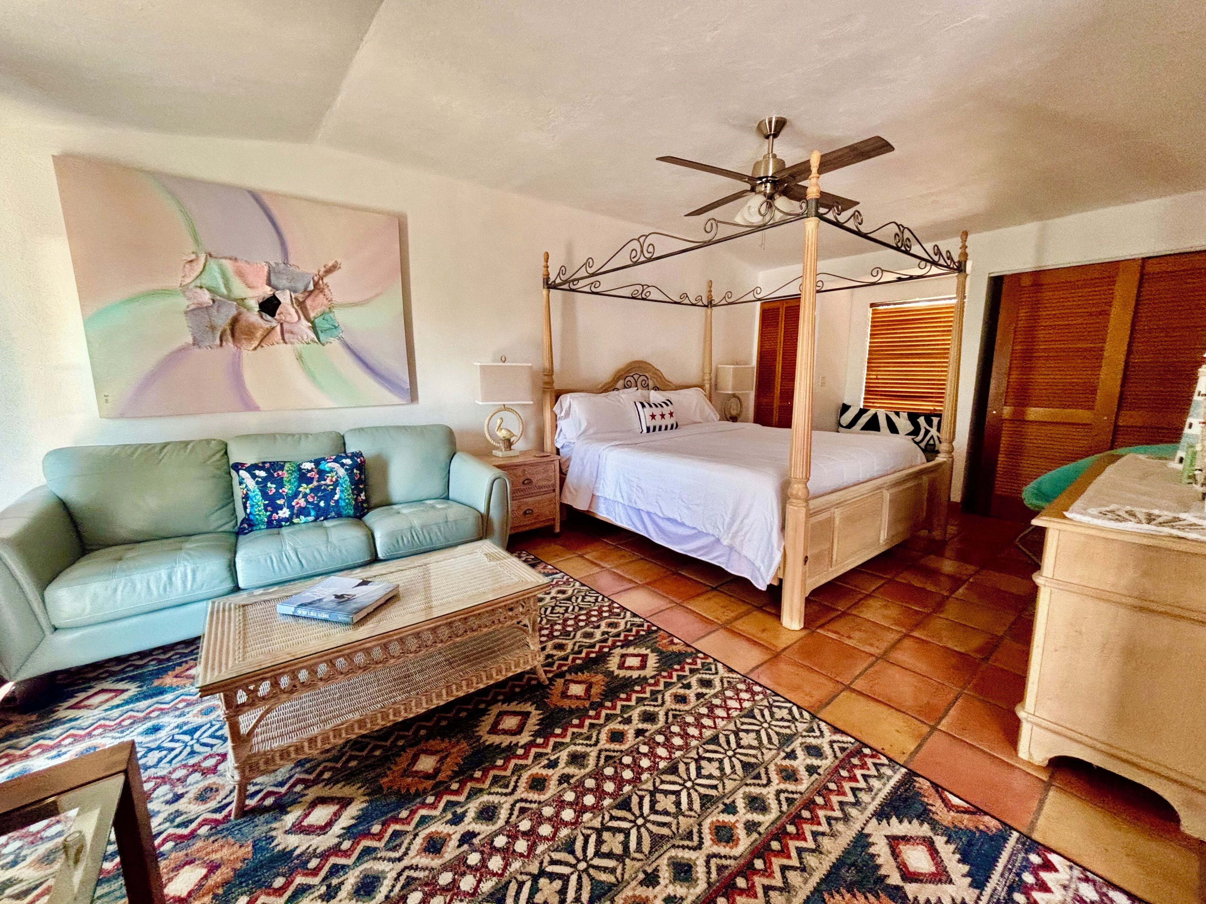 A four-poster bed with a ceiling fan, a sofa with a floral cushion, a patterned rug, and a painting on the wall.