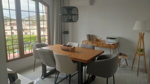 Een eetgedeelte met een houten tafel, zes stoelen, een hanglamp, uitzicht op groen en gebouwen door grote ramen, en een klein bureau met een printer en een lamp.