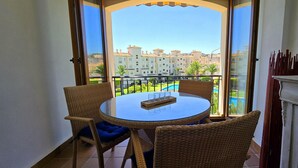 Een balkon met een ronde tafel en rieten stoelen, met uitzicht op een zwembad en woongebouwen.