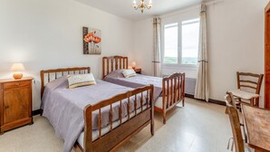 A bedroom with two wooden beds, a wooden nightstand, a wooden chair, a wooden table, a chandelier, a window with curtains, and a wall-mounted painting. - Gîte La Motette Drôme Provençale (Rousset les Vignes)