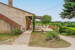 A building with a brick exterior, a covered patio with outdoor furniture, and a view of a lush green landscape.