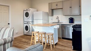 Una cocina compacta con lavadora y secadora integradas, estufa y fregadero. Hay dos taburetes de madera y una silla a rayas.