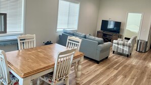 Una sala de estar con una mesa de comedor de madera, dos sillas blancas, un sofá gris y un televisor sobre un mueble de madera.