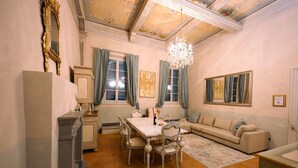 A dining area with a table, chairs, and a chandelier. There is a sofa and a window with curtains. - Avignonesi - Tuscan Palace (Montepulciano)