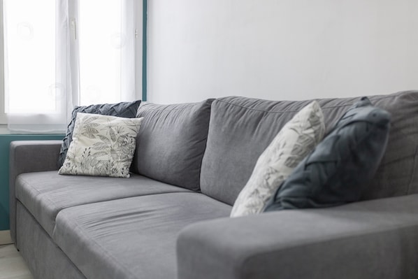 Un sofá gris con almohadones estampados, una ventana con cortinas traslúcidas y una pared de acento color aguamarina.