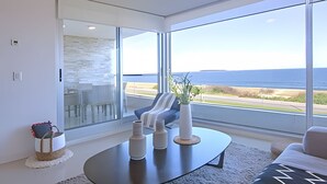 Een moderne woonkamer met een grote glazen deur die leidt naar een balkon, een ronde salontafel en uitzicht op de oceaan.