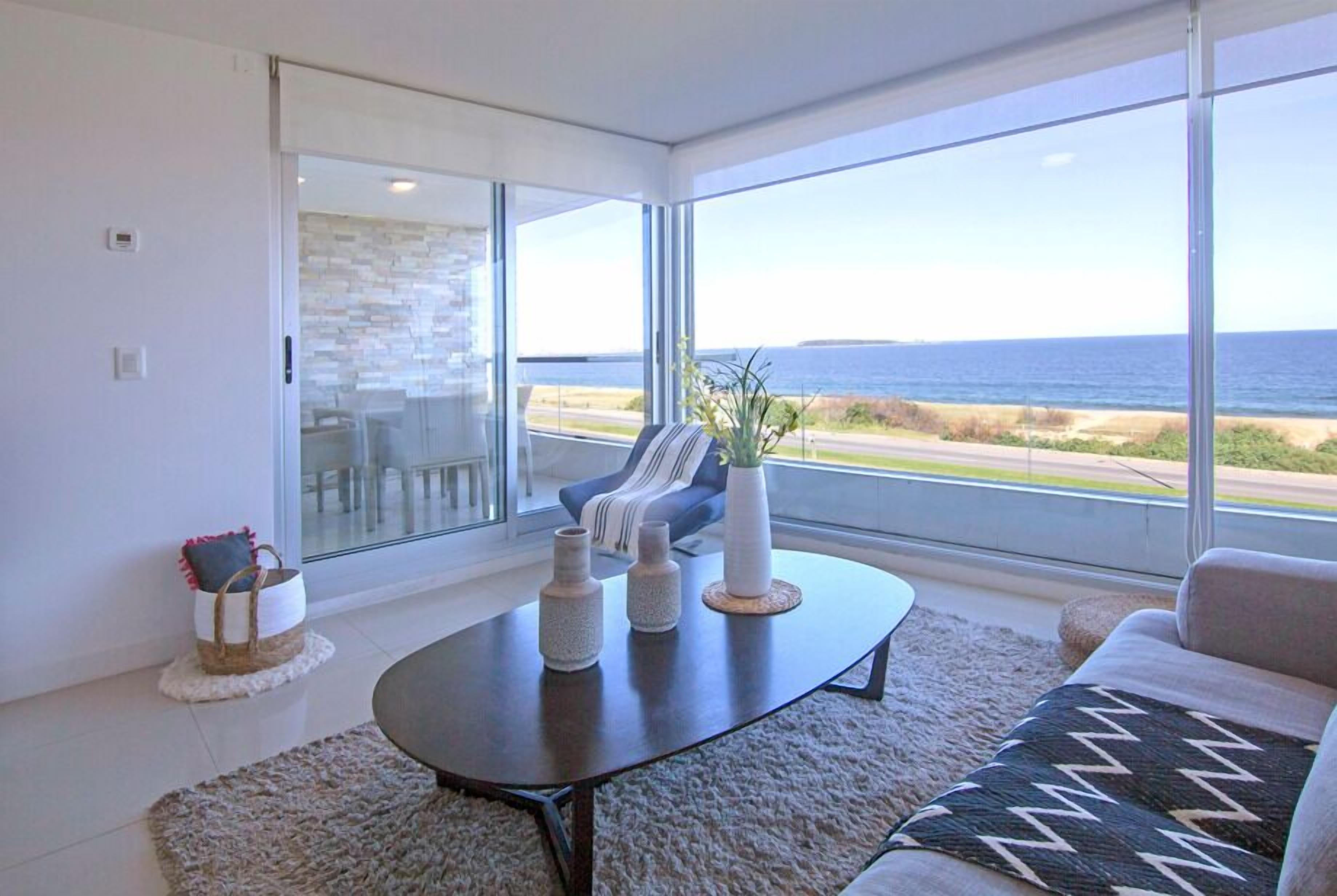 A modern living room with a large glass door leading to a balcony, a dark coffee table, and a view of the ocean.