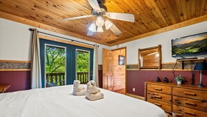 Ein Schlafzimmer mit einem großen Bett, Holzbalkendecke, Deckenventilator, Fernseher, Kommode und Blick ins Freie.