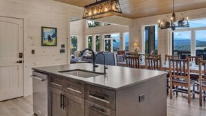 A modern kitchen with a central island, wooden dining area, and large windows offering views of the outdoors. - New! Loaded Luxe w/Pool*Theater*Game Room*Views! (Sevierville)