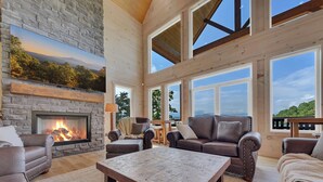 A spacious living room with a stone fireplace, leather sofas, and large windows offering a view of the outdoors. - New! Loaded Luxe w/Pool*Theater*Game Room*Views! (Sevierville)