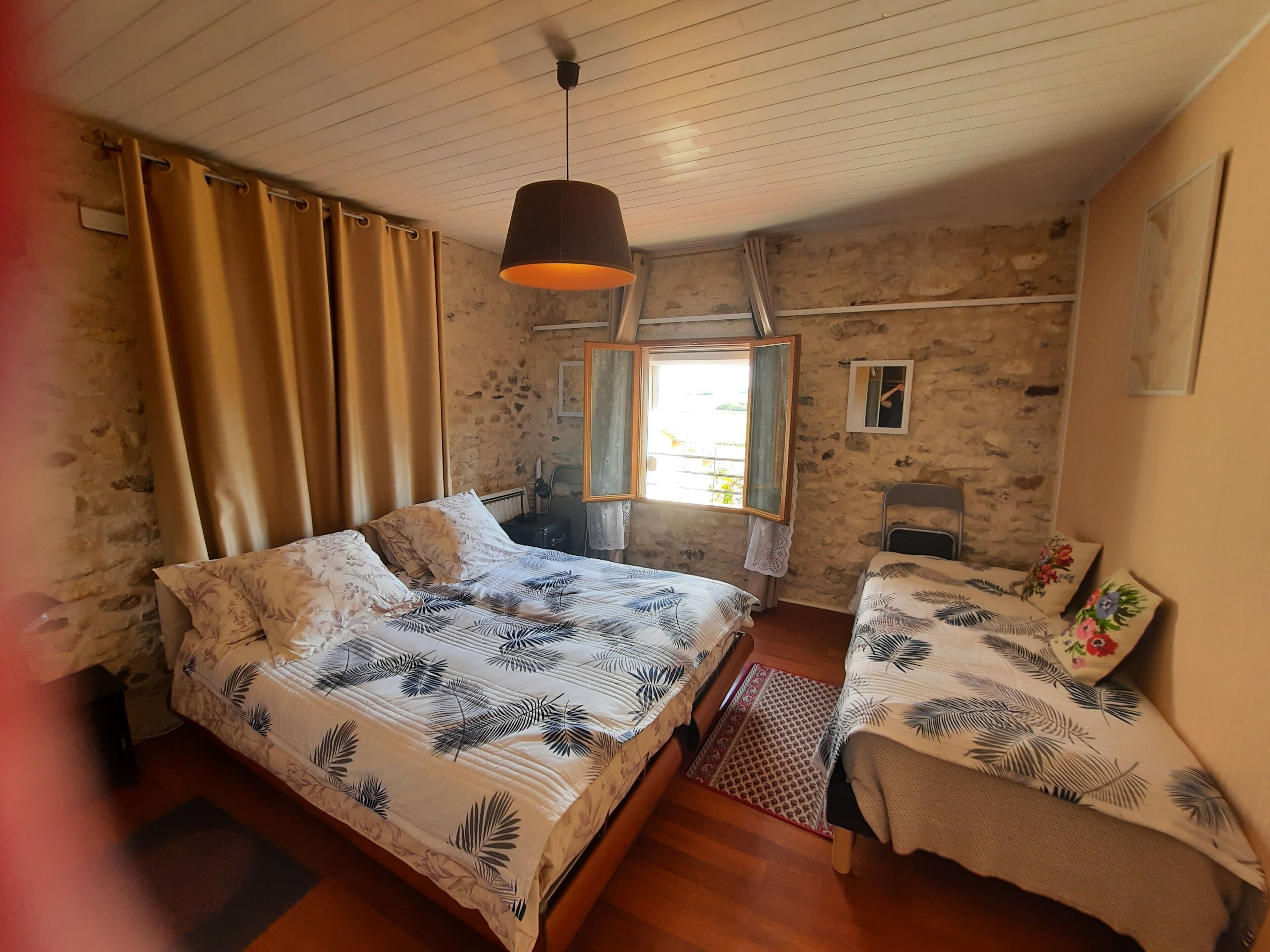 A bedroom with a large bed, a smaller bed, a window with curtains, a hanging light fixture, and a stone wall.