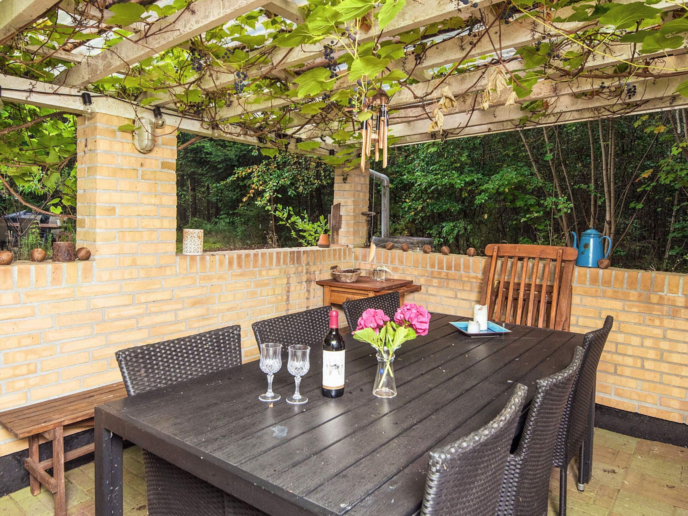 An outdoor patio with a brick wall, a table set with wine glasses and a bottle, and a pergola with vines.