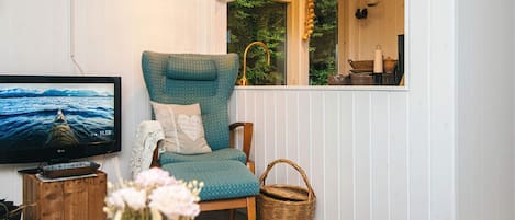 A cozy living room with a television, a comfortable chair, a wooden side table, a vase with flowers, and a window with a view of greenery.