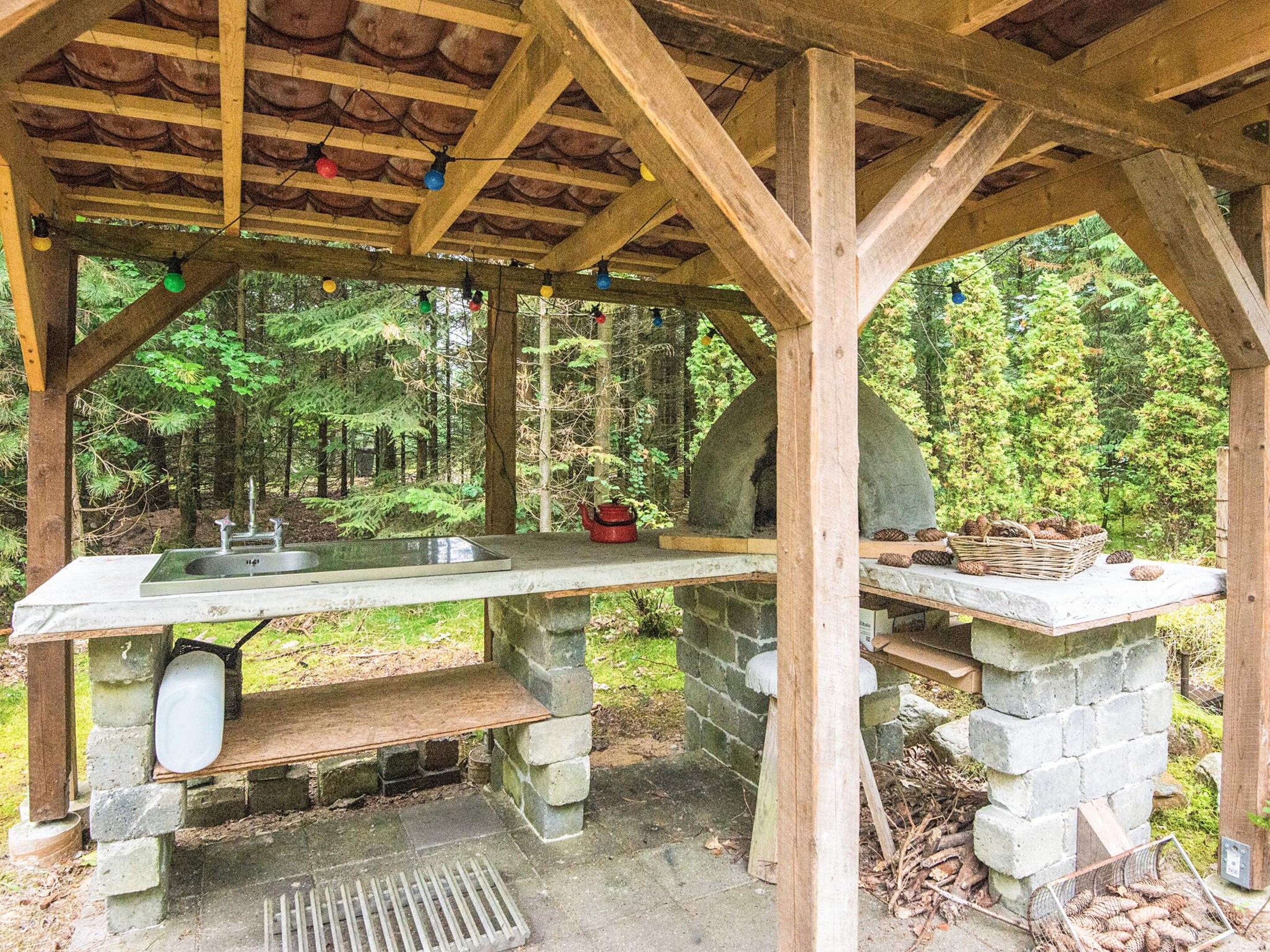 Una cocina rústica al aire libre con un lavabo de piedra, un hervidor rojo y una cesta de piñas.