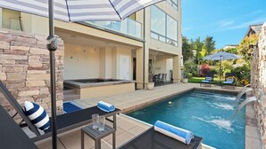 Un espace piscine extérieur moderne, agrémenté d’un mur en pierre, de chaises longues et d’une table avec un parasol à rayures.