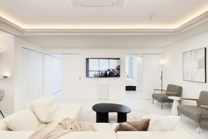 A modern living room with a sofa, coffee table, and armchairs, a flat-screen TV mounted on the wall, and a view into another room through an open door.