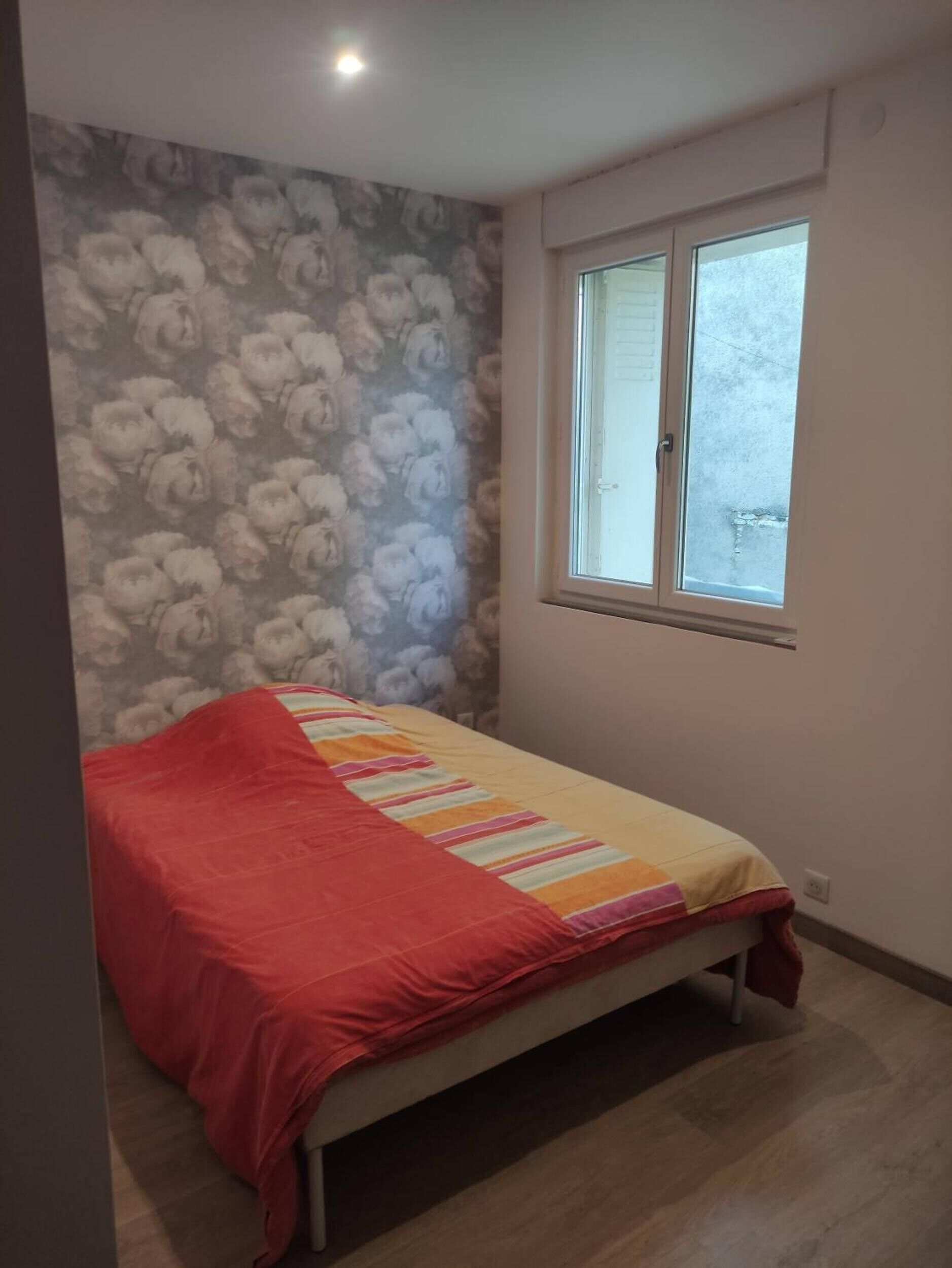 A single bed with a striped bedspread, a window with blinds, and a floral-patterned wall.