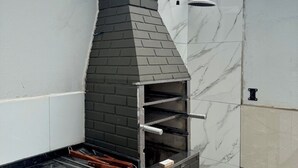 A modern outdoor kitchen with a brick chimney, a black countertop, and a glass enclosure. - Beach house in a gated community. Enclosed area with barbecue | Small pets allowed (Caraguatatuba)