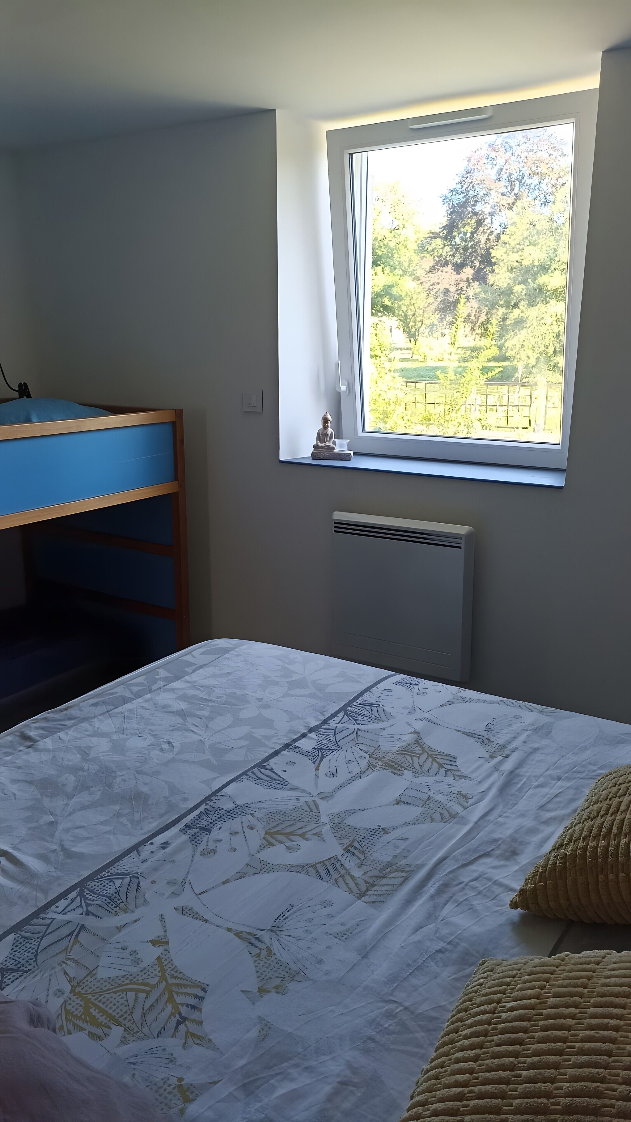 Un dormitorio con cama, ventana con vistas a follaje, radiador y escritorio azul.