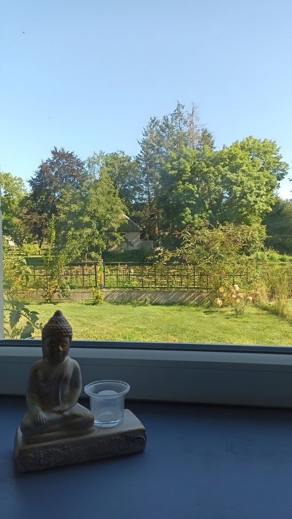 A Buddha statue on a table by a window overlooking a garden with trees and a fence.
