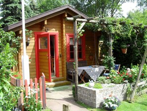 Una cabaña de madera con puertas y ventanas rojas, una jardinera de piedra y una mesa con mantel.