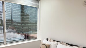 Ein modernes Schlafzimmer mit einem großen Bett, einem Fenster mit Jalousien und Blick auf eine Stadtlandschaft.