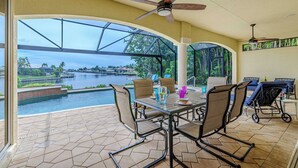 A covered patio with a table and chairs, overlooking a swimming pool and a view of a body of water with buildings in the distance. - Southern Exposure - 4bed/3bath - Sands Area - Stars Vacation Rentals (Cape Coral)