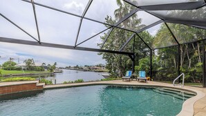 An indoor swimming pool with clear water, surrounded by a glass enclosure, and a view of a canal with houses and palm trees in the background. - Southern Exposure - 4bed/3bath - Sands Area - Stars Vacation Rentals (Cape Coral)