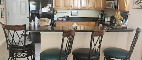 A kitchen with wooden cabinets, a black refrigerator, a microwave, and a coffee maker. There are four black bar stools with green cushions.