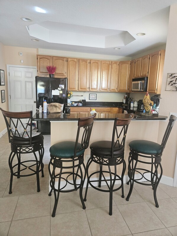 A kitchen with wooden cabinets, a black refrigerator, a microwave, and a coffee maker. There are four black bar stools with green cushions.