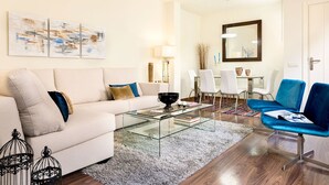 Una sala moderna con un sofá blanco, una mesa de centro de vidrio, una silla azul y un comedor con sillas y una mesa blancos.