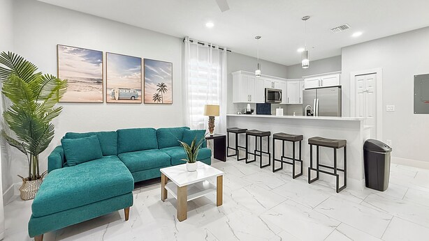 A modern living room with a teal sofa, a white coffee table, and a kitchen area with white cabinets and a stainless steel refrigerator.