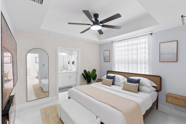 Ein modernes Schlafzimmer mit einem großen Bett, einem Deckenventilator, einem Spiegel, einem Badezimmer mit weißem Waschtisch und einem großen Fenster mit durchsichtigen Vorhängen.