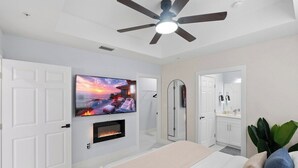 A modern bedroom with a TV mounted above a fireplace, a ceiling fan, and a walk-in closet.