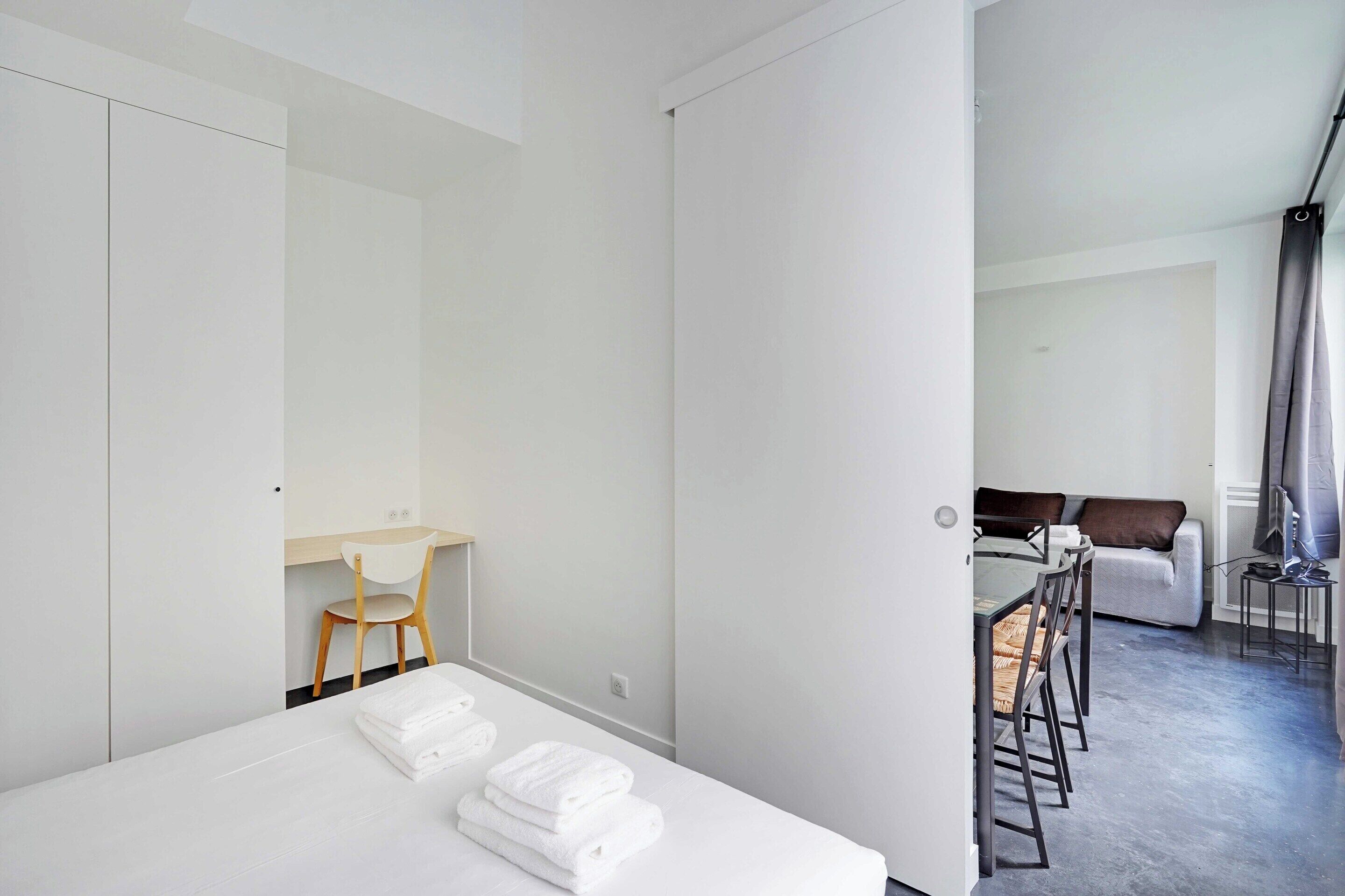 A modern hotel room with a neatly made bed, a desk with a chair, and a sofa in the background.