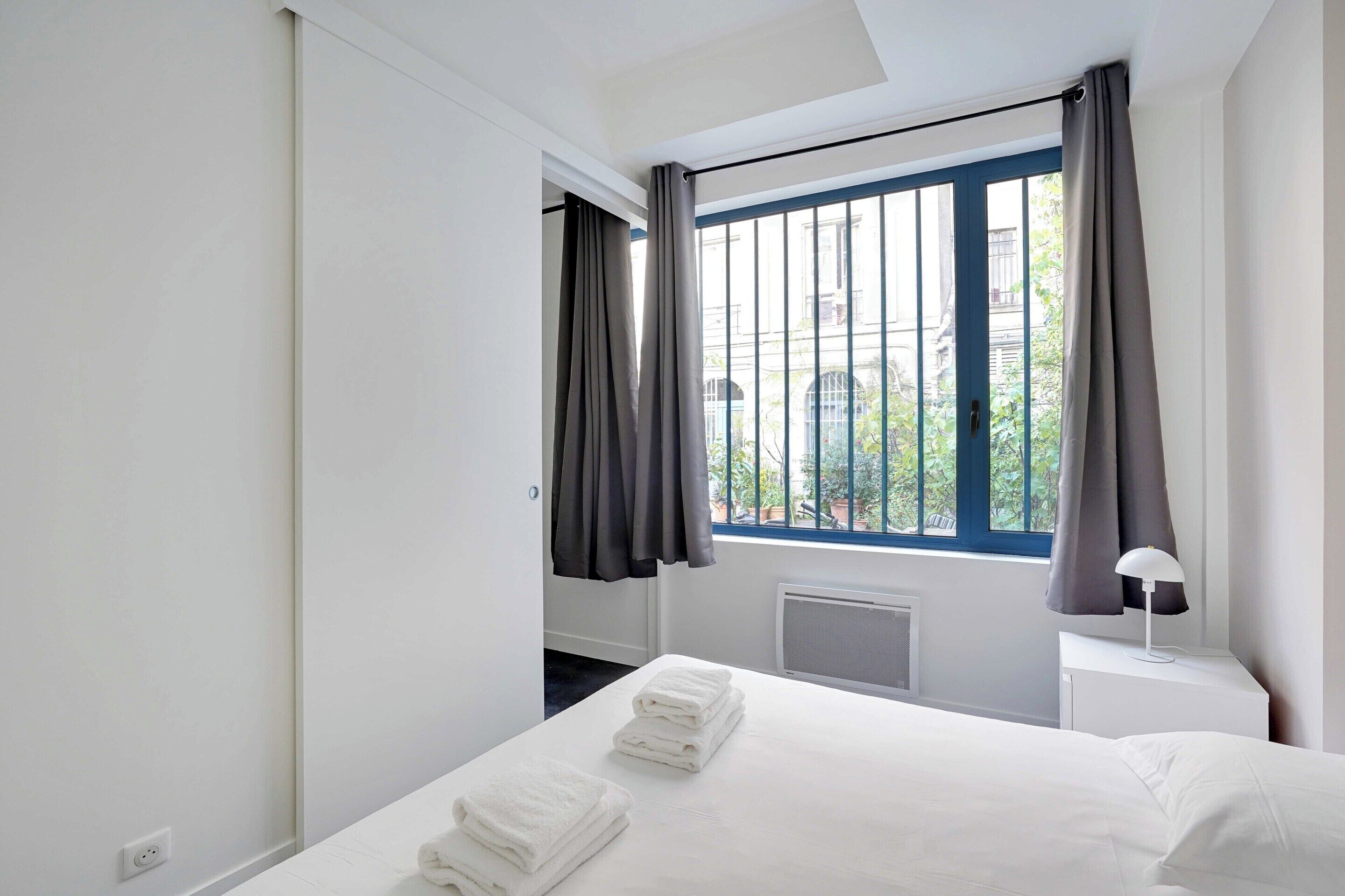 Une chambre d’hôtel moderne avec un grand lit, du linge de lit blanc, une fenêtre avec des rideaux, une table de chevet avec une lampe et une vue sur l’extérieur.