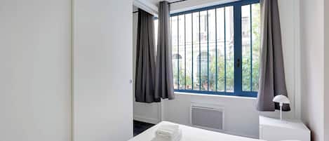 A modern hotel room with a large bed, white linens, a window with curtains, a bedside table with a lamp, and a view of the outside.